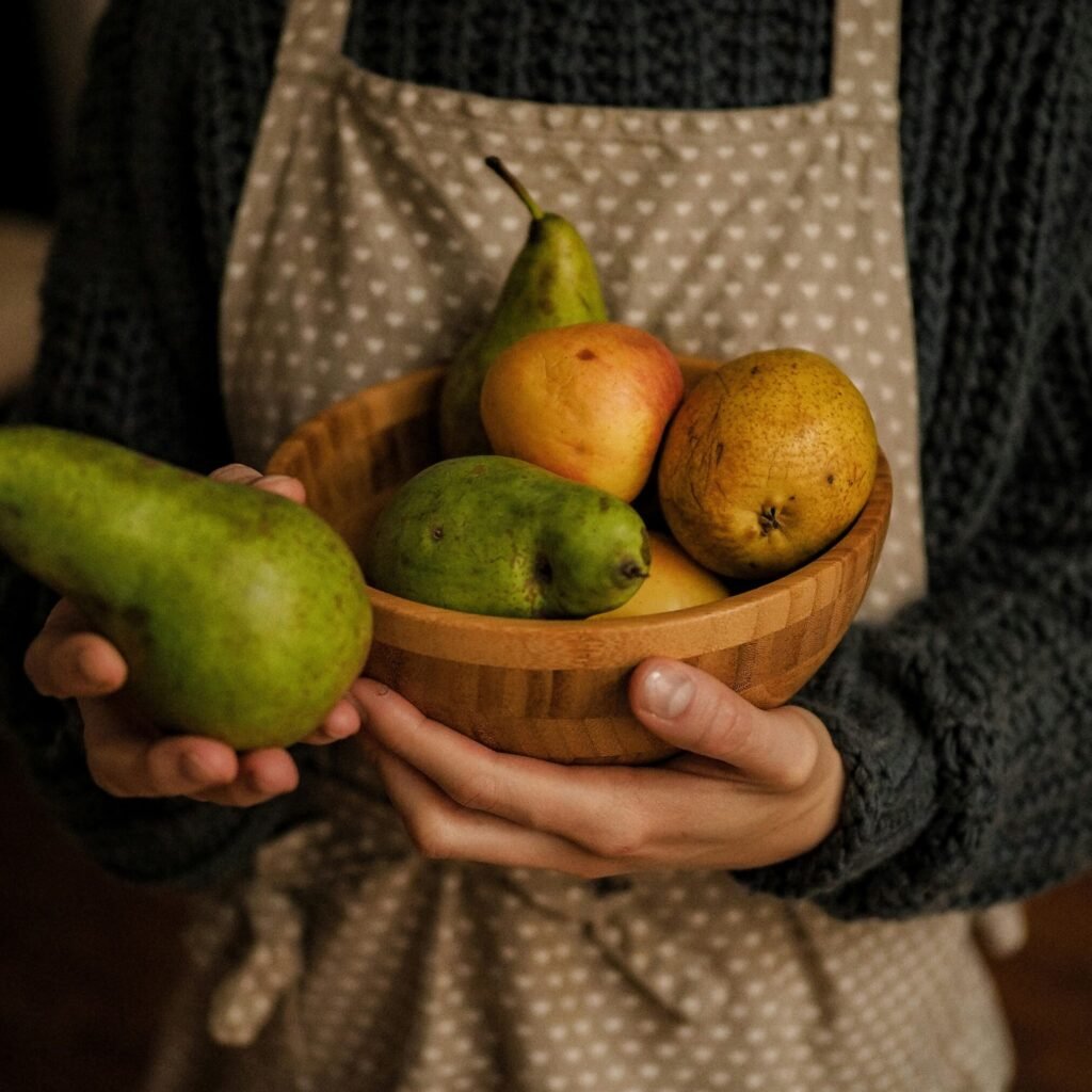 Hands holding a wooden bowl filled with pears and apples, wearing an apron.